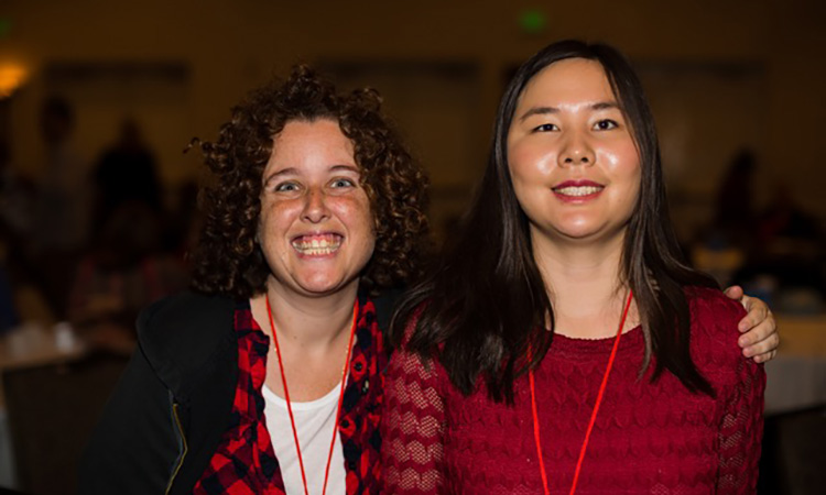 Two women smile, one Asian and one white and both wearing red pose with their arms around each other