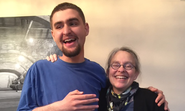 A young man with short brown hair and a small beard stands with his arm around a woman, his mother, while both of them laugh. He is a full head taller than her.