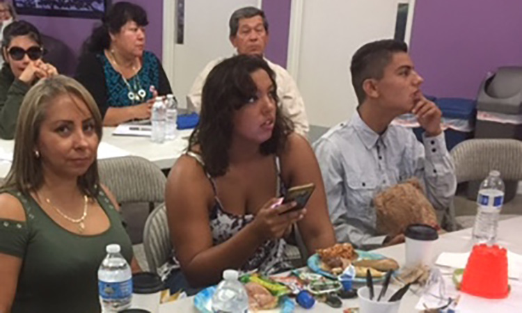 Two women and a man with a disability sit at a table with food, looking off toward a presentation