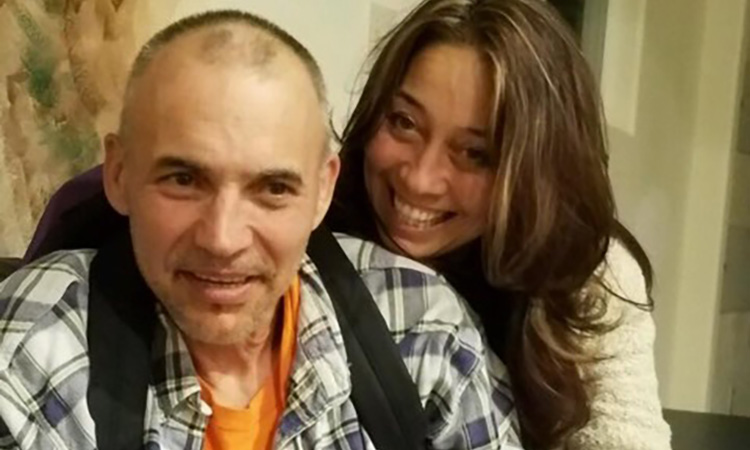 A man with a buzz cut wearing a backpack smiles while a woman with long brown hair hugs him from behind and smiles widely