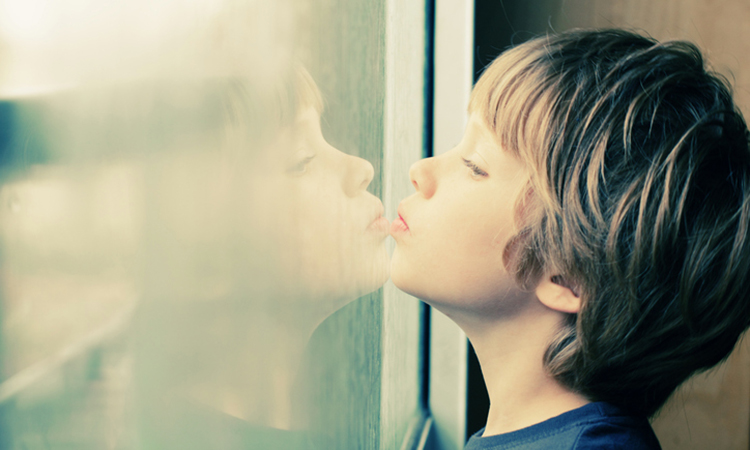 A young white boy with blonde hair looks out a window, his face reflected back at him