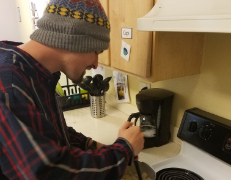 A man with a small beard wearing a dark blue and red flannel shirt and a gray beanie stands in front of a stovetop stirring food in a pot