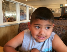 A Latino boy with short dark hair looks off to the side while sitting in a diner with a small smile. 