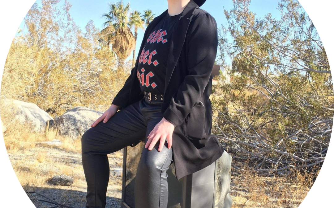 Full-body image of Ed Hirtzel, a white butch with glasses and short blonde hair wearing all black, including a black wide-brimmed hat and black, spiked mask. Ed is sitting on a TV in 3/4 profile view, in the desert near her home.