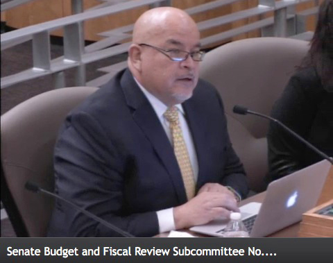 DVU Vice President Fernando Gomez sits in a suit at a table with a laptop in front of him, testifying at a legislative hearing. Text on image reads: Senate Budget and Fiscal Review Subcommittee No…