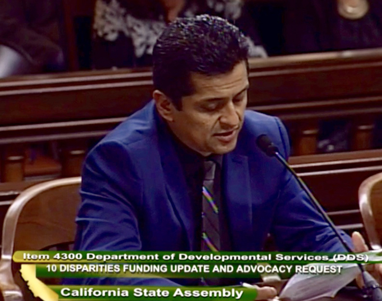 A Latino man in a blue suit testifies in a courthouse, holding papers. Text on image reads: Iten 4300 Department of Developmental Services (DDS) 10 DISPARITIES FUNDING UPDATE AND ADVOCACY REQUEST. California State Assembly.