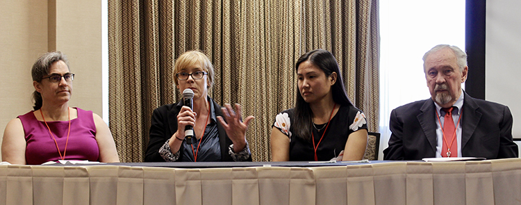 Two white women, an Asian woman, and white man sit on a panel at a conference, with the second woman speaking into a microphone