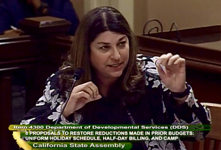DVU President Judy Mark testifies from a witness table, one hand raised in front of her, her face focused. Text along bottom of image reads: Item 4300 Department of Developmental Services (DDS) 8 Proposals to Restore Reductions Made in Prior Budgets: Uniform Holiday Schedule, Half-Day Billing, and Camp. California State Assembly