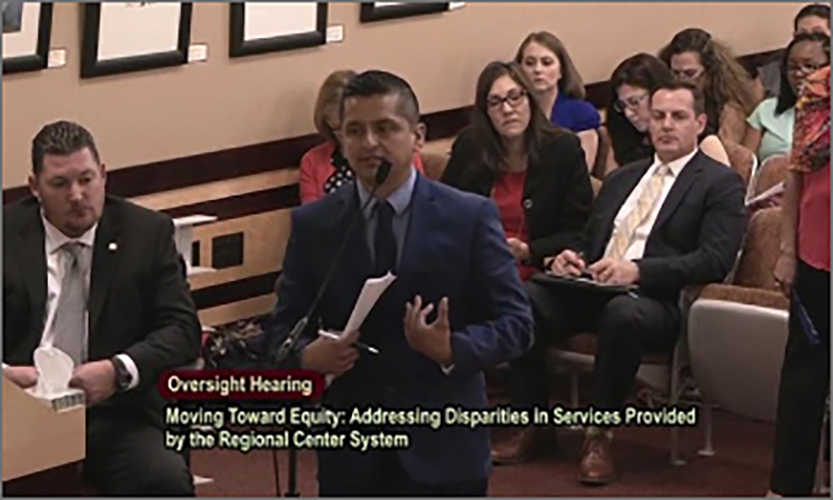 A Latino man speaks at a microphone, wearing a suit and holding papers. People sit in rows behind him. Text on image reads: Oversight Hearing. Moving Toward Equity: Addressing Disparities in Services Provided by the Regional Center System