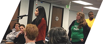 A Latina woman with dark hair stands in front of a microphone, a line behind her and an audience around her, all facing the same direction