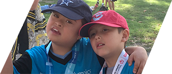 Two Latino boys, one with Down syndrome, and both wearing uniforms and caps, have their arms around each other