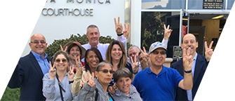 DVU board members and self-advocates pose in a big group in front of the Santa Monica Courthouse, all smiling and holding up the victory sign with their fingers in a V