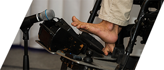 A man’s foot is tapping on a communication device attached to the base of his wheelchair, which is next to a microphone