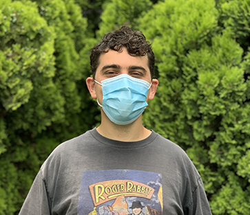 A young white man with short brown curly hair stands in front of a hedge wearing a gray Roger Rabbit t-shirt and a blue surgical mask. His eyes are squinted in a smile. 
