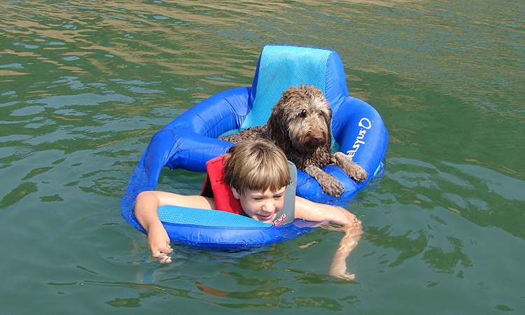 A young boy with blonde hair and a life vest shares a flotation device with a brown dog