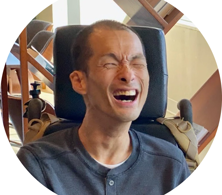 Image of Tim Jin, an Asian man with very short black hair, smiling widely at the camera. He is seated in a wheelchair and wearing a dark gray henley.
