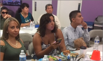 Two Latina women and a Latino man with a disability sit at a table with food, looking off toward a presentation